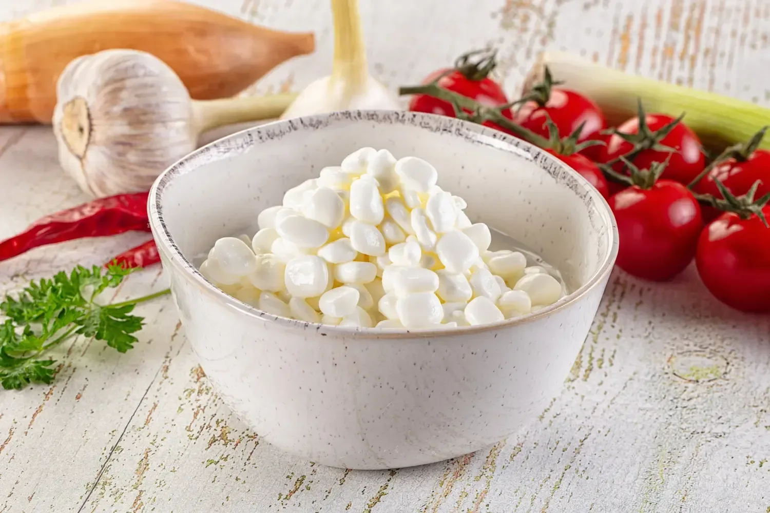 Un bol de cottage cheese aux grains blancs, entouré d'ail, de tomates cerises, de céleri, de carotte, de piment et de persil