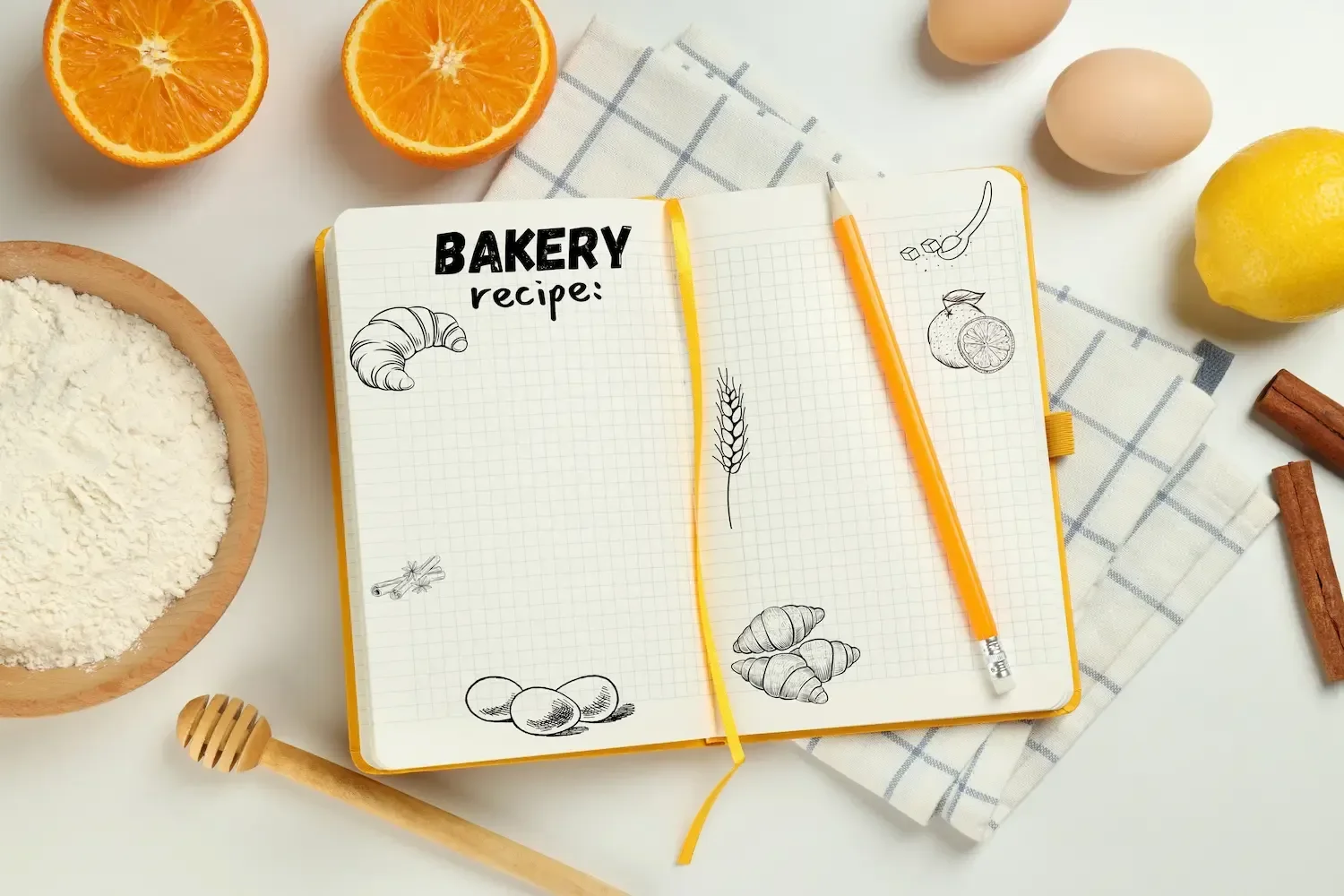 Carnet de recettes ouvert sur une page quadrillée avec un crayon, entouré de farine, d'œufs, d'oranges et de bâtons de cannelle